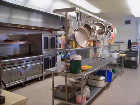 Industrial kitchen with hanging pots, stainless steel appliances, and prep stations in a care facility.