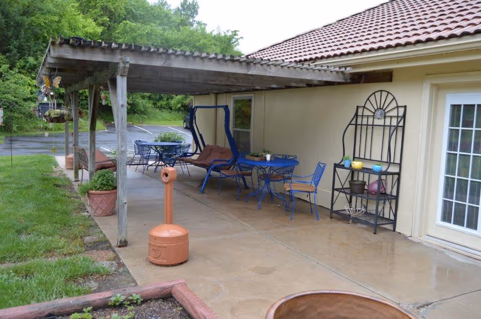 Covered outdoor patio with blue metal tables and chairs, a swinging bench, potted plants, and a decorative shelving unit beside a building.