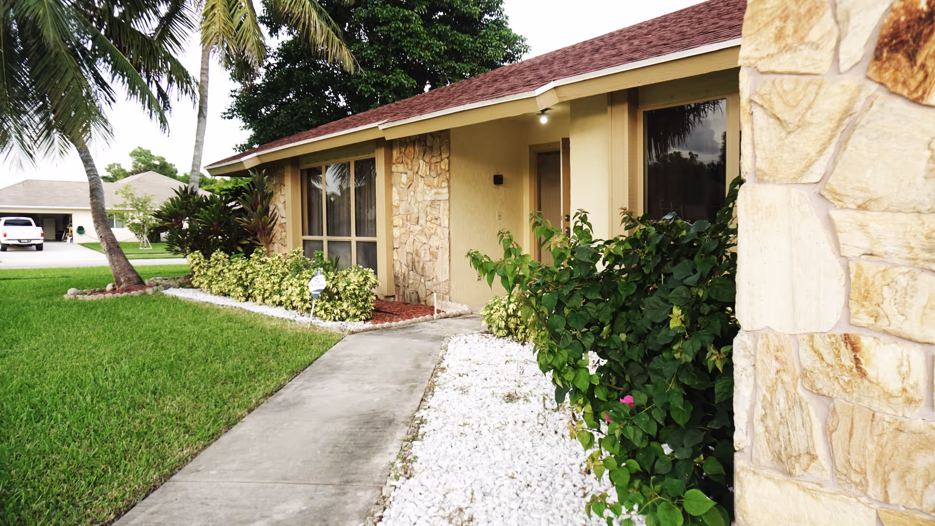 Exterior view of a single-story assisted living facility building with stone and beige walls, large windows, a concrete walkway leading to the entrance, surrounded by green grass, bushes, and palm trees.