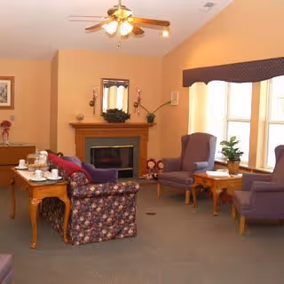 A cozy living room with a floral patterned sofa, two purple armchairs, wooden side tables, a fireplace with a mirror above it, and large windows with a valance allowing natural light to fill the room.