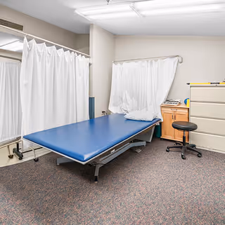 Small medical exam room with a blue padded treatment table, white privacy curtains, a rolling stool, and storage cabinets.