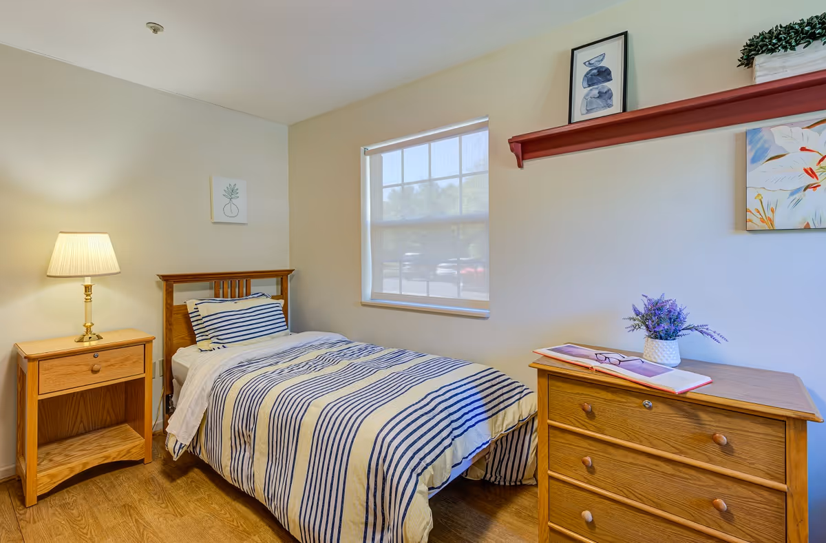A small bedroom with a single bed covered in a white and blue striped comforter and matching pillow. To the left of the bed is a wooden nightstand with a lamp on it. On the right side of the room is a wooden dresser with an open book, a pair of glasses, and a small potted plant on top. Above the dresser is a red wall shelf holding a framed picture and a small plant. The room has a window with a white blind partially drawn, letting in natural light. The walls are light-colored and the floor is wooden.