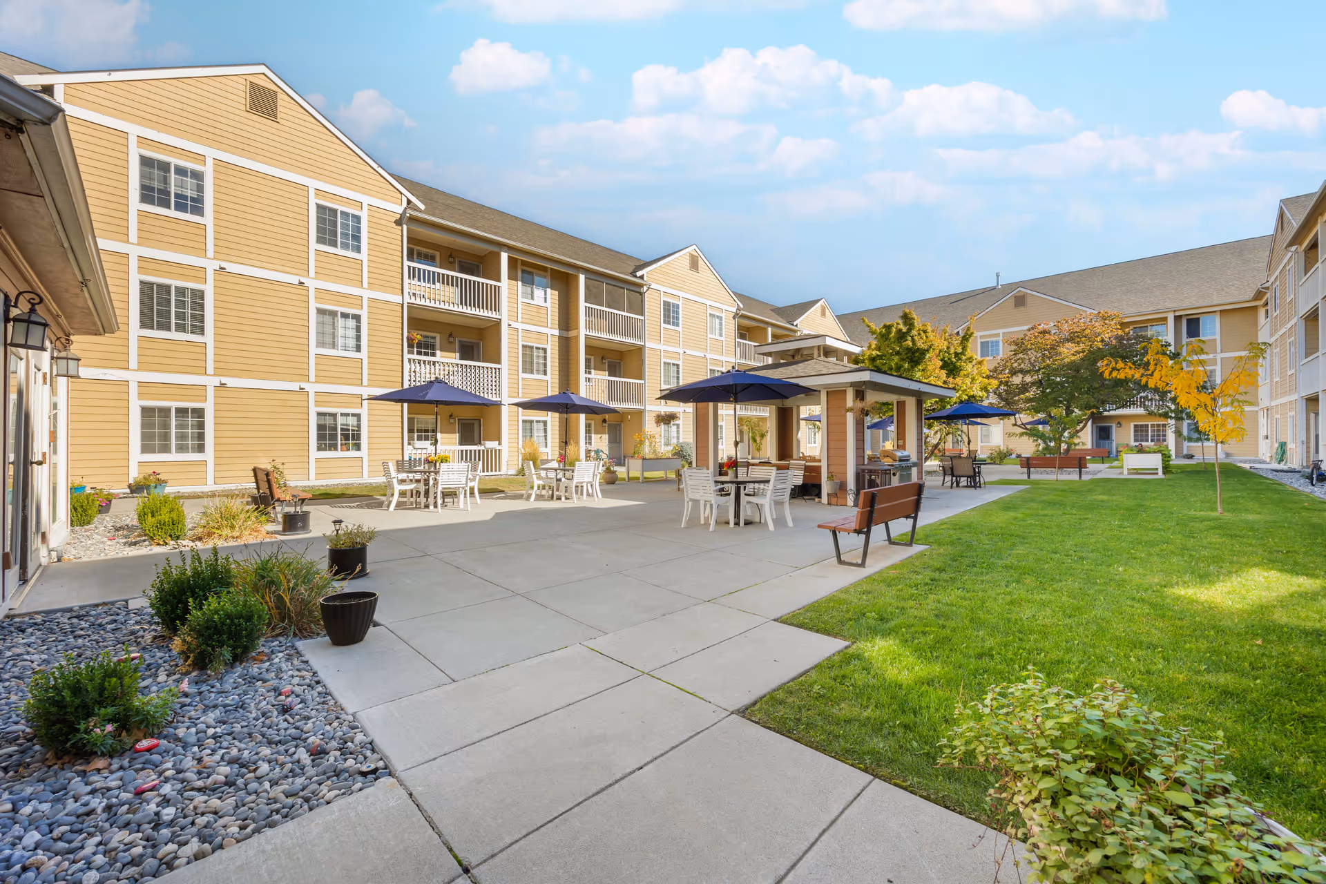 Outdoor courtyard area of a senior living facility with beige multi-story buildings surrounding a paved patio. The patio has tables with blue umbrellas, chairs, benches, and a small covered seating area. There is a well-maintained green lawn and some trees and shrubs around the patio under a partly cloudy sky.
