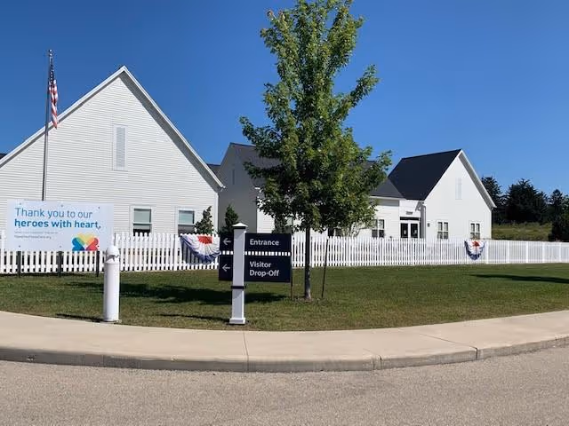 Exterior view of a white building with a white picket fence and a tree in front. There is a sign indicating entrance and visitor drop-off. A banner on the fence reads 'Thank you to our heroes with heart.' The sky is clear and blue.