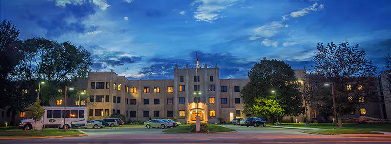 Exterior view of Buehler Home building at dusk with illuminated windows, parked cars, trees, and a partly cloudy sky.