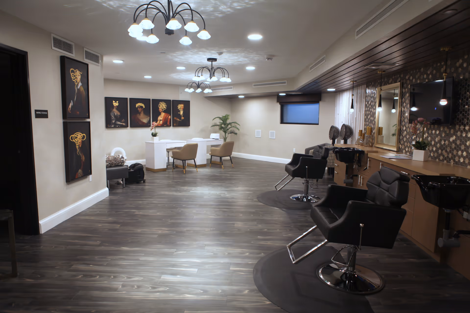 Interior of a modern salon area in a senior living facility with black salon chairs, wash basins, a wall-mounted TV, decorative wall art, and a small seating area with two beige chairs and a white table. The floor is dark wood, and the ceiling has multiple light fixtures.