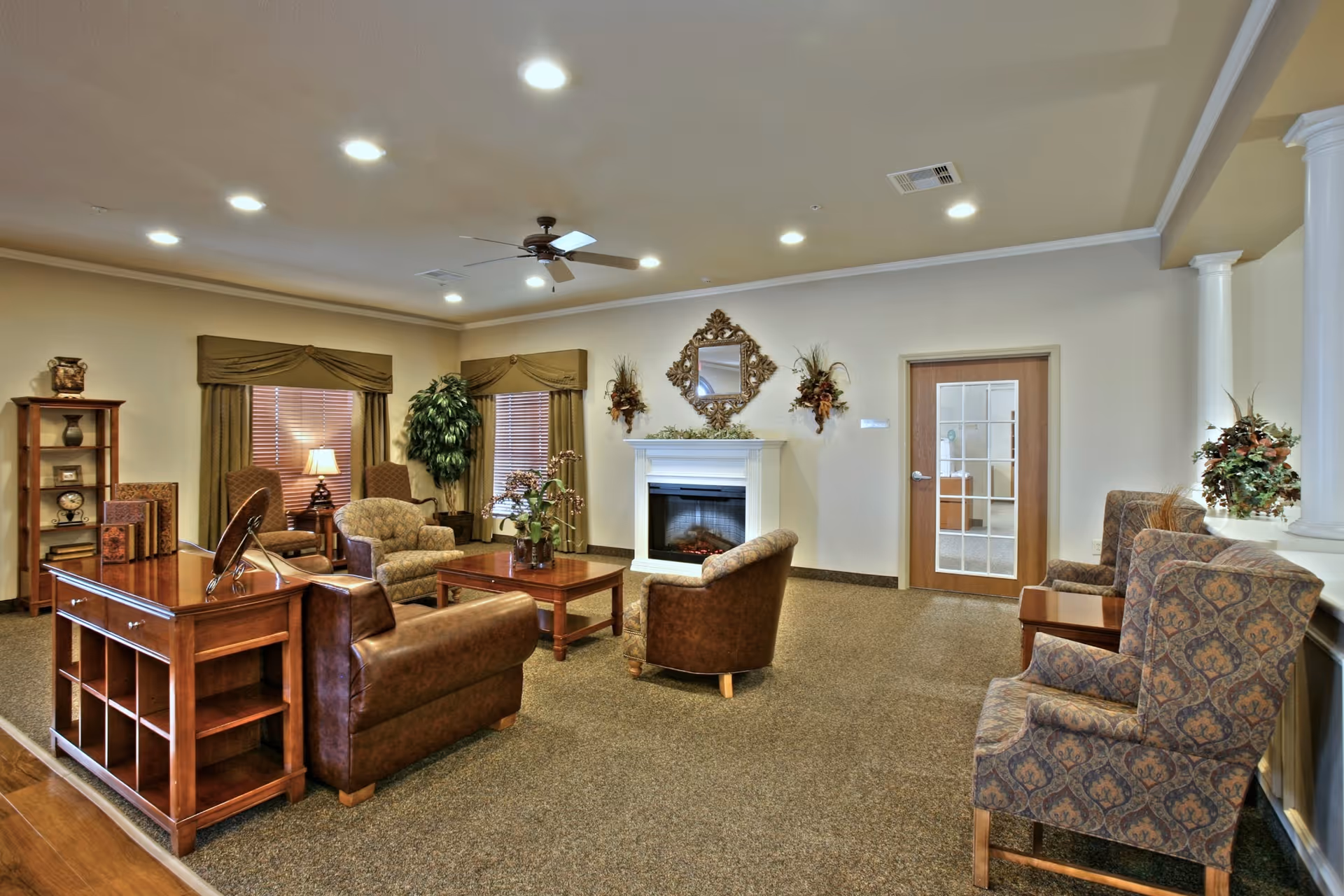 A cozy living room area in a senior living facility featuring a fireplace with a decorative mirror and floral arrangements above it. The room has multiple upholstered chairs and sofas arranged around a wooden coffee table. There are two windows with brown curtains, a ceiling fan, recessed lighting, and a wooden door with glass panels. The decor includes plants and a wooden shelving unit with decorative items.