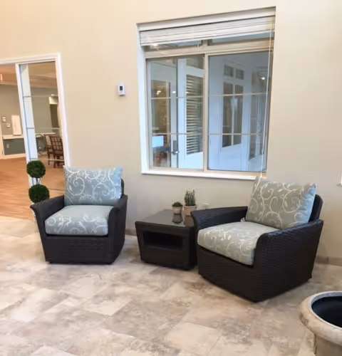 Two cushioned wicker armchairs with light blue patterned cushions are placed on a tiled floor with a small dark wicker side table between them holding two small potted plants. Behind the chairs is a large window with white blinds and a beige wall. To the left, an open doorway leads to another room with wooden flooring and additional seating visible.