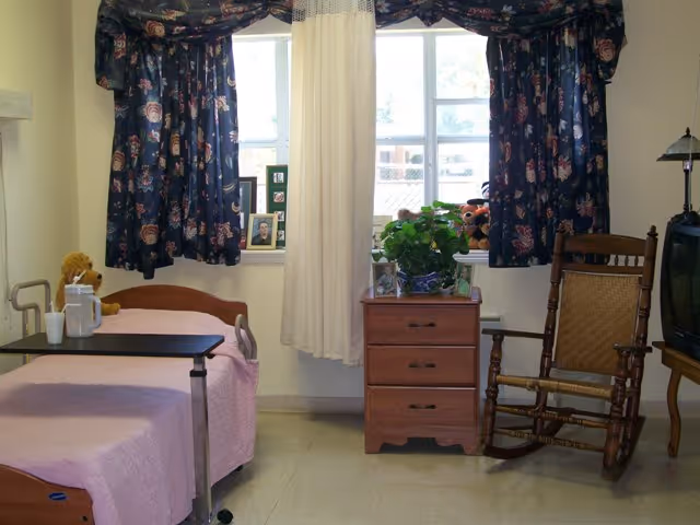 A small nursing home bedroom with a single bed covered in a pink blanket, a bedside table with a plant and framed photos, a wooden rocking chair, a television on a stand, and a window with floral curtains and a sheer white curtain in the center.