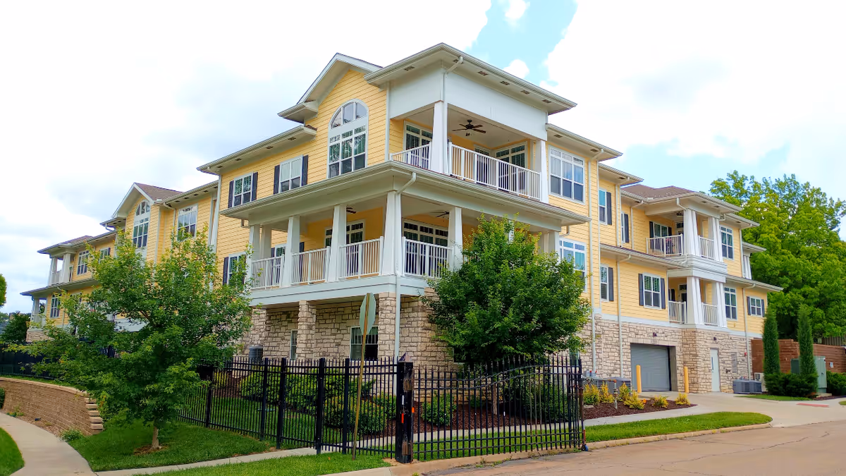 Exterior view of a three-story senior living facility building with yellow siding and stone accents. The building features multiple balconies with white railings, surrounded by green trees and landscaping. A black metal fence encloses part of the property, and a paved road runs alongside the building under a partly cloudy sky.