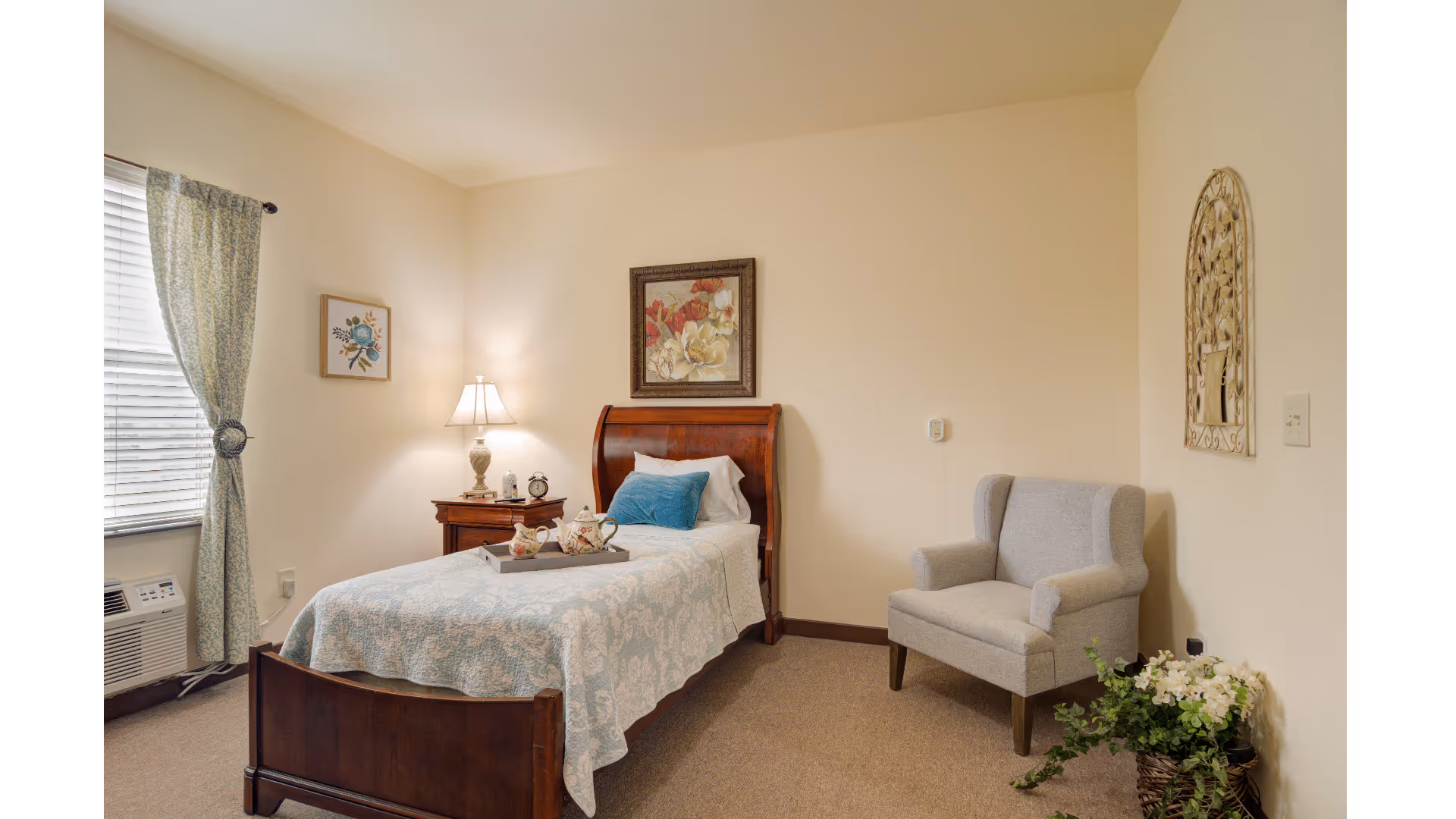 A cozy senior living bedroom with a single wooden bed covered in a light blue patterned quilt and a blue pillow. A wooden nightstand with a lamp, clock, and decorative items is beside the bed. There is a gray upholstered armchair in the corner next to a basket of white flowers. The room has beige walls, a window with patterned curtains, and framed floral artwork on the walls.