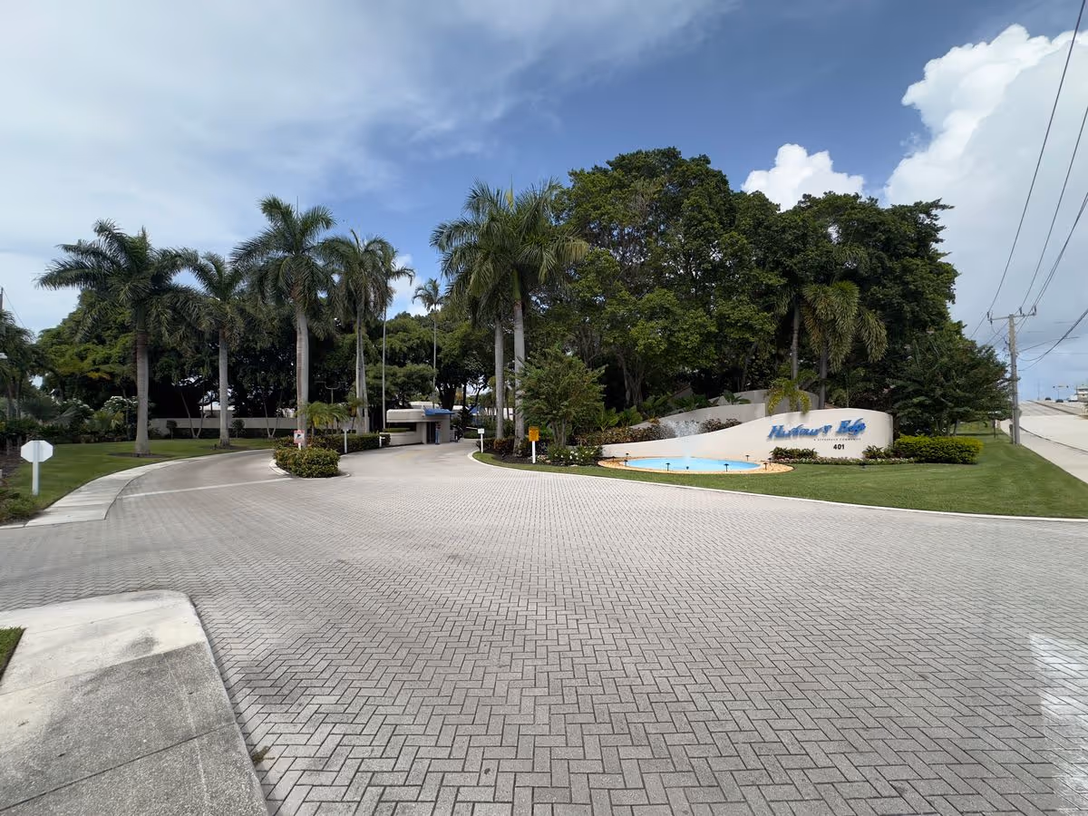 Paved driveway leading to the landscaped entrance and sign of a residential complex flanked by palm trees and greenery under a partly cloudy sky.