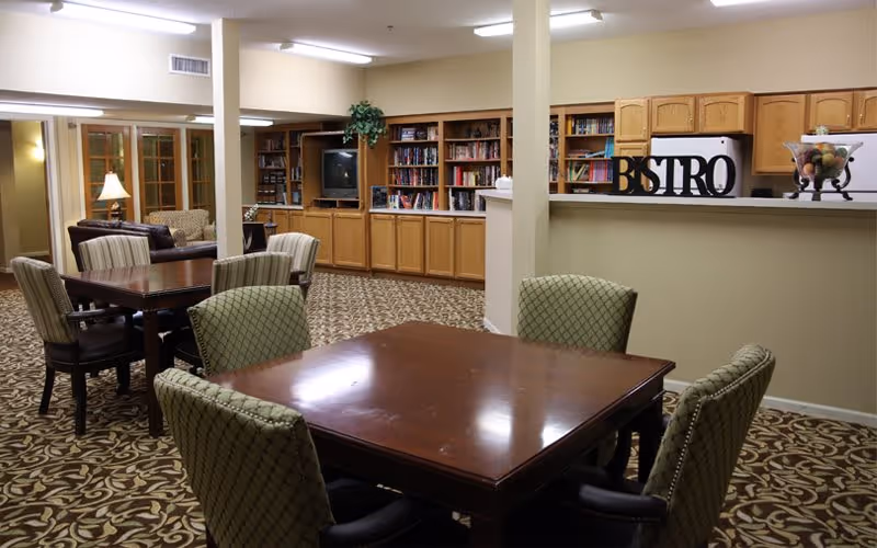 A cozy common area with multiple wooden tables and cushioned chairs arranged on a patterned carpet. In the background, there is a built-in wooden bookshelf filled with books and a television. To the right, a counter with a decorative sign that reads 'BISTRO' and a bowl of fruit is visible. The room is softly lit with ceiling lights and a table lamp near a glass door.
