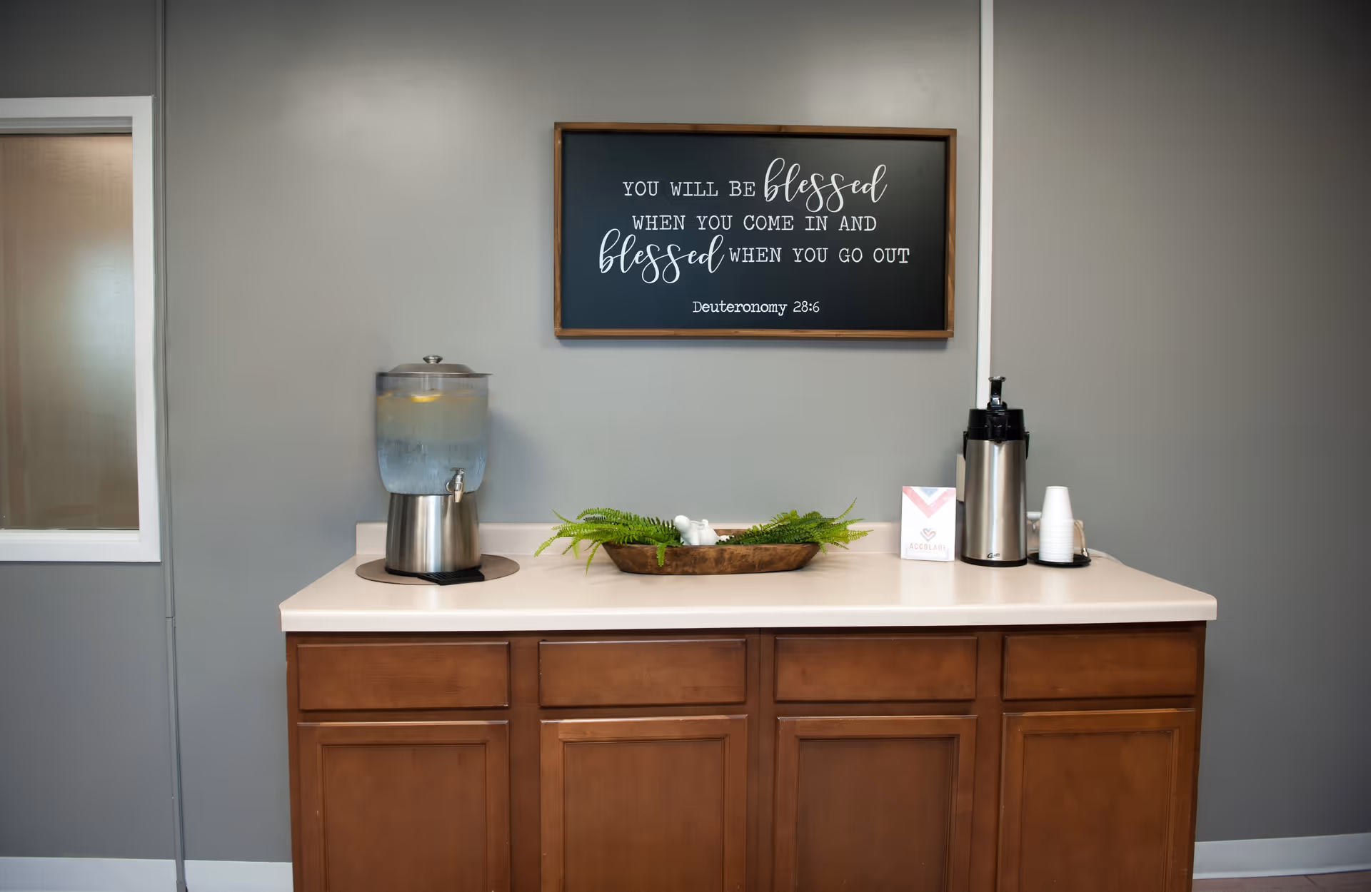 A countertop with a water dispenser, a decorative wooden tray with green ferns and a small white bird figurine, a coffee dispenser with stacked white cups, and a small sign with the Accolade logo. Above the countertop is a framed sign with the quote: 'You will be blessed when you come in and blessed when you go out. Deuteronomy 28:6'. The wall behind is painted gray and there is a window to the left.
