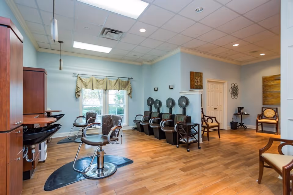 Interior view of a salon area in a senior living facility with light blue walls, wooden flooring, and ceiling lights. The room features salon chairs with black sinks, hair drying stations with multiple hooded dryers, and several wooden chairs along the walls. A window with blinds and a valance allows natural light into the room.