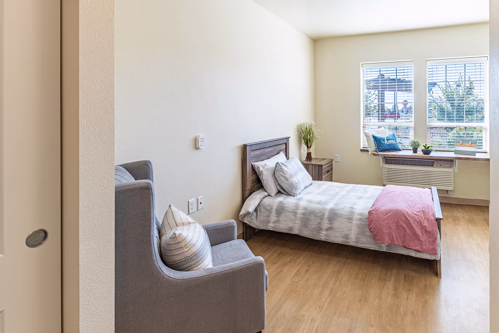 A bright and clean bedroom with a single bed made with light gray and white bedding and a pink throw blanket. Next to the bed is a wooden nightstand with a vase of decorative grass. A gray armchair with a striped pillow is positioned near the bed. The room has light-colored walls and wood flooring. Two large windows with blinds let in natural light, and a window ledge holds a blue pillow with the word 'home', two small potted plants, and a decorative box.