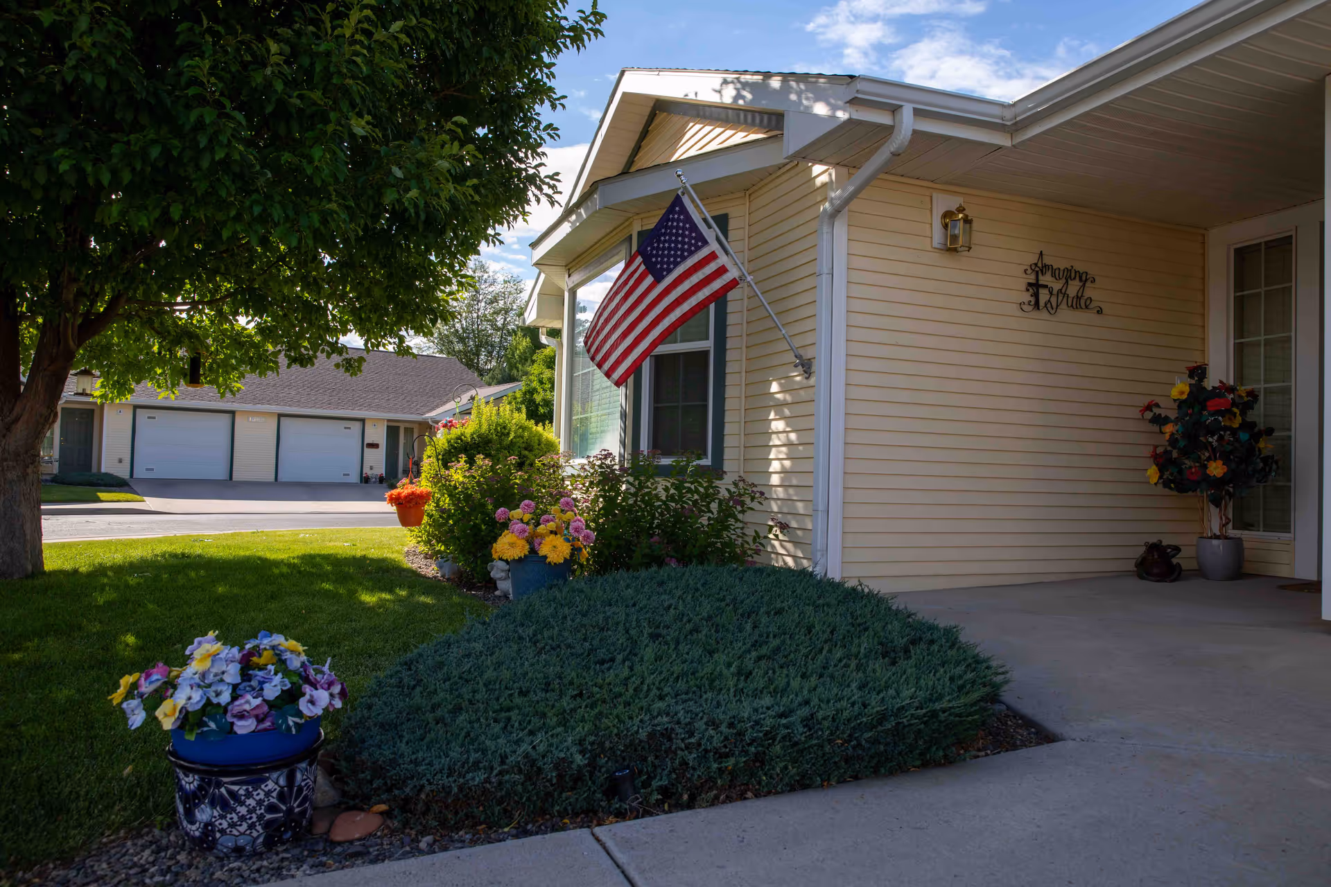 Exterior view of a residential building with beige siding, an American flag mounted near a window, and a porch area decorated with potted flowers and a plant. There is a tree and well-maintained lawn in the foreground, and a neighboring building with garages is visible in the background under a partly cloudy sky.