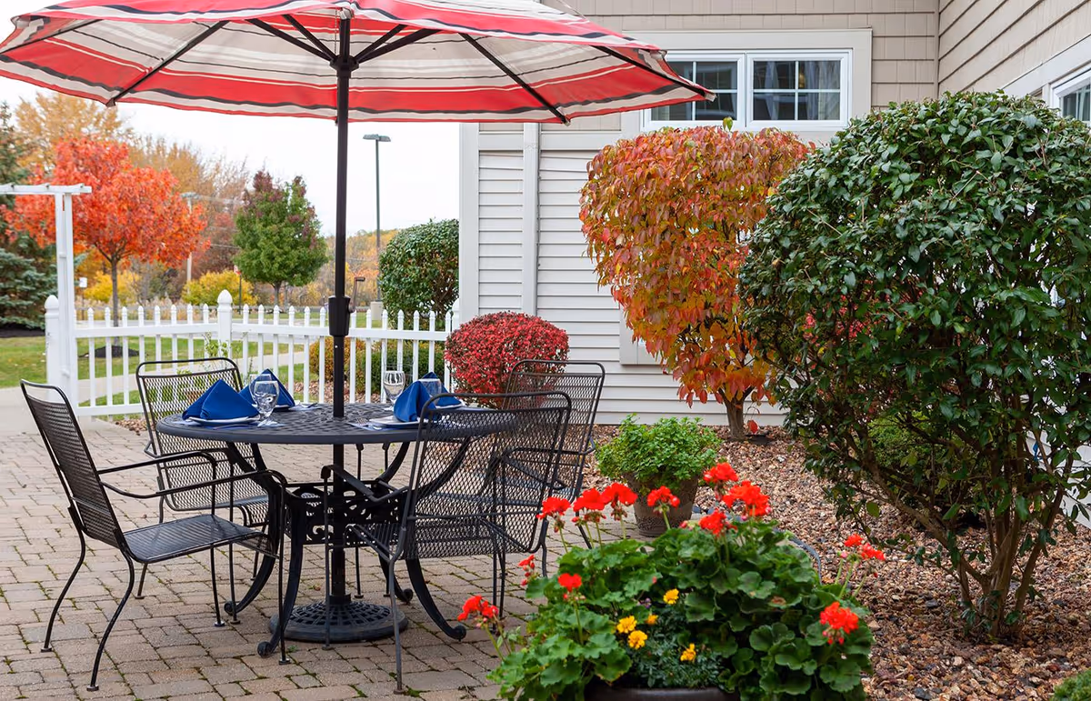 Outdoor patio area with a round black metal table and four matching chairs under a large red and white striped umbrella. The table is set with blue napkins and glassware. Surrounding the patio are various bushes and plants, including red and yellow flowers, with a white picket fence and trees with autumn foliage in the background.