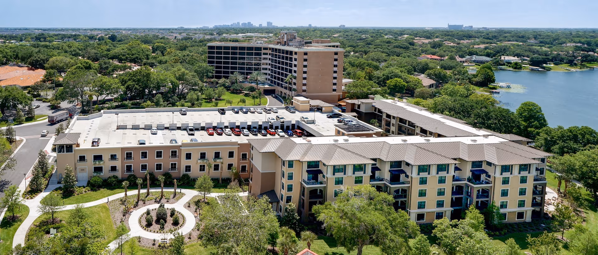 Aerial view of Westminster Winter Park senior living facility showing multiple connected buildings with beige and light brown exteriors, surrounded by green trees and landscaped gardens. There is a circular garden feature in the foreground and a lake visible to the right side of the image under a clear sky.