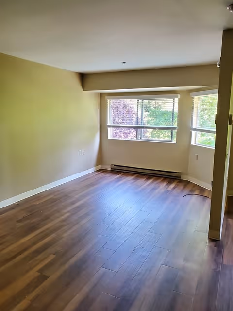 Empty room with wooden flooring, beige walls, and a large window with blinds showing green and purple foliage outside.