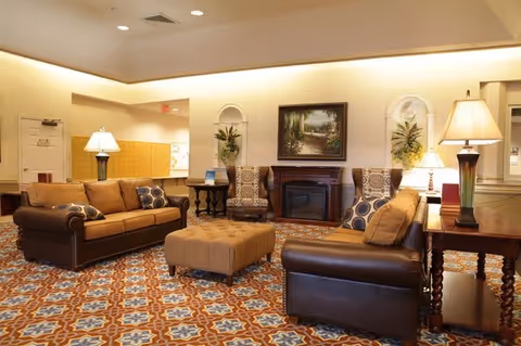 A cozy senior living facility lounge area with two brown leather sofas, a patterned ottoman, two patterned armchairs, side tables with lamps, a fireplace, and a framed painting on the wall. The room has warm lighting and a decorative patterned carpet.