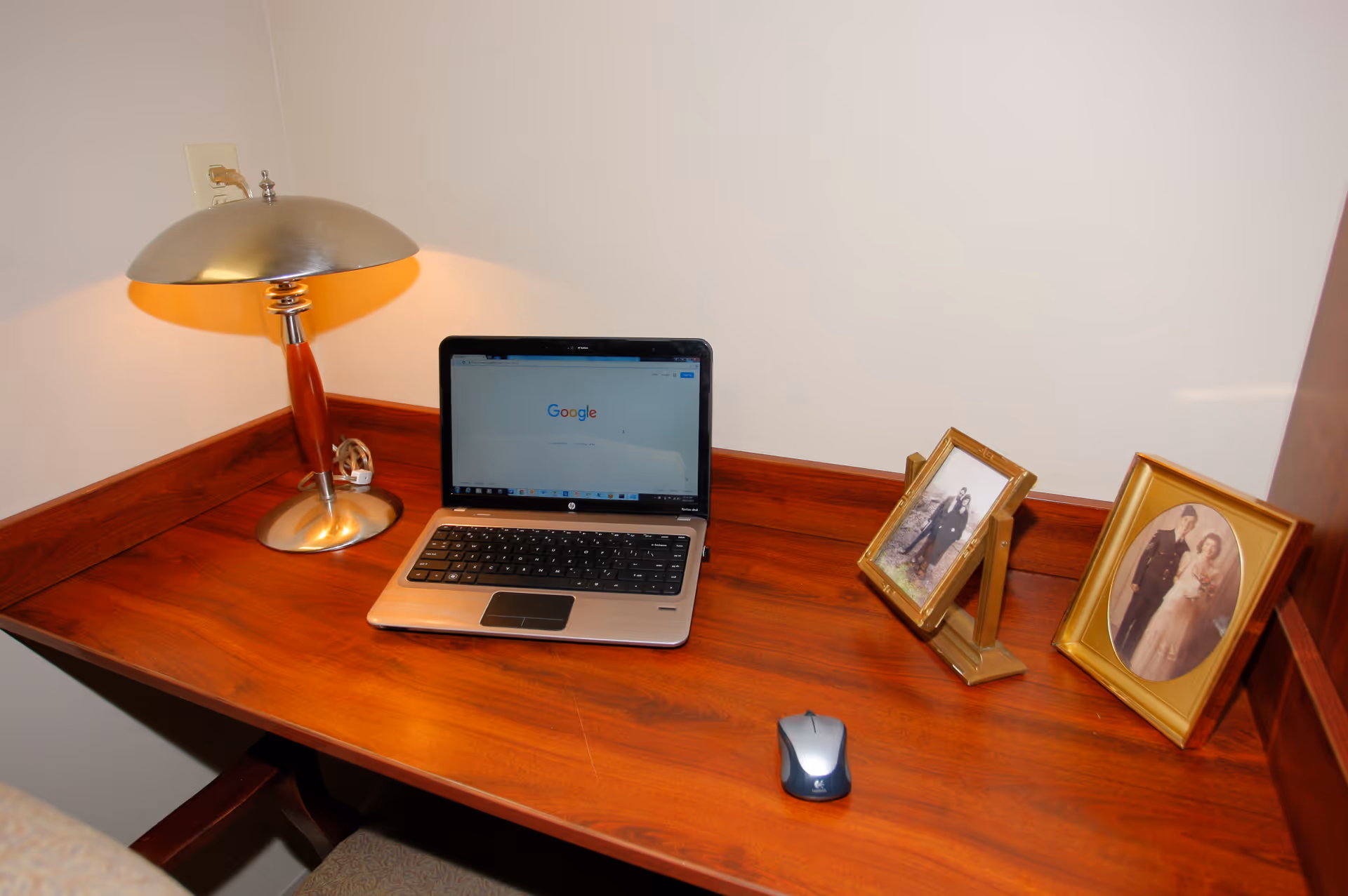 A wooden desk with a silver and wood table lamp, an open laptop displaying the Google homepage, a wireless mouse, and two framed photographs on the right side.