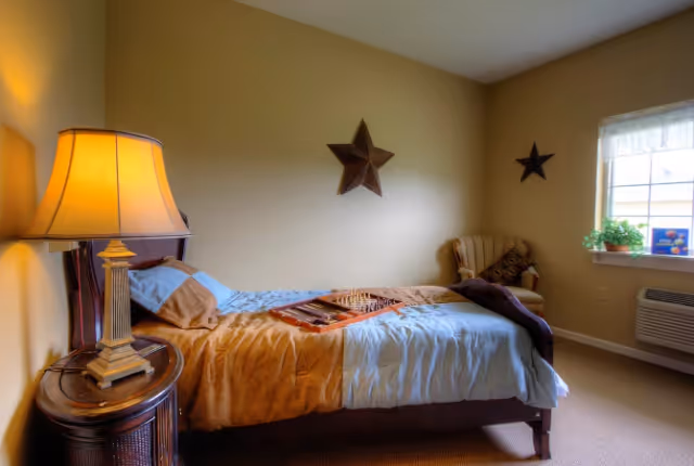 Small furnished bedroom with a single bed, bedside lamp on a nightstand, decorative star wall hangings, an armchair by the window, and a board game on the bed.