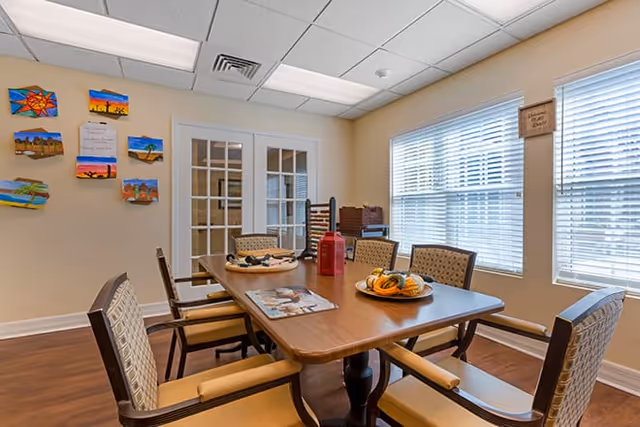 A dining room with a rectangular wooden table surrounded by chairs, place settings, and large windows with blinds.