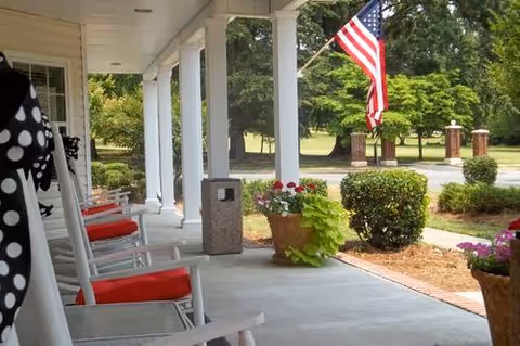 Covered front porch with white rocking chairs with red cushions, potted plants, and an American flag by the driveway.
