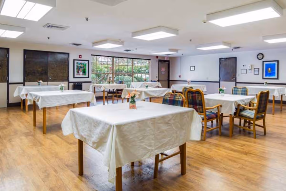 A spacious dining room with several tables covered in white tablecloths, each adorned with a small flower arrangement. The room has wooden flooring, large windows letting in natural light, and a few framed pictures on the walls. There are cushioned chairs with plaid upholstery around some of the tables.