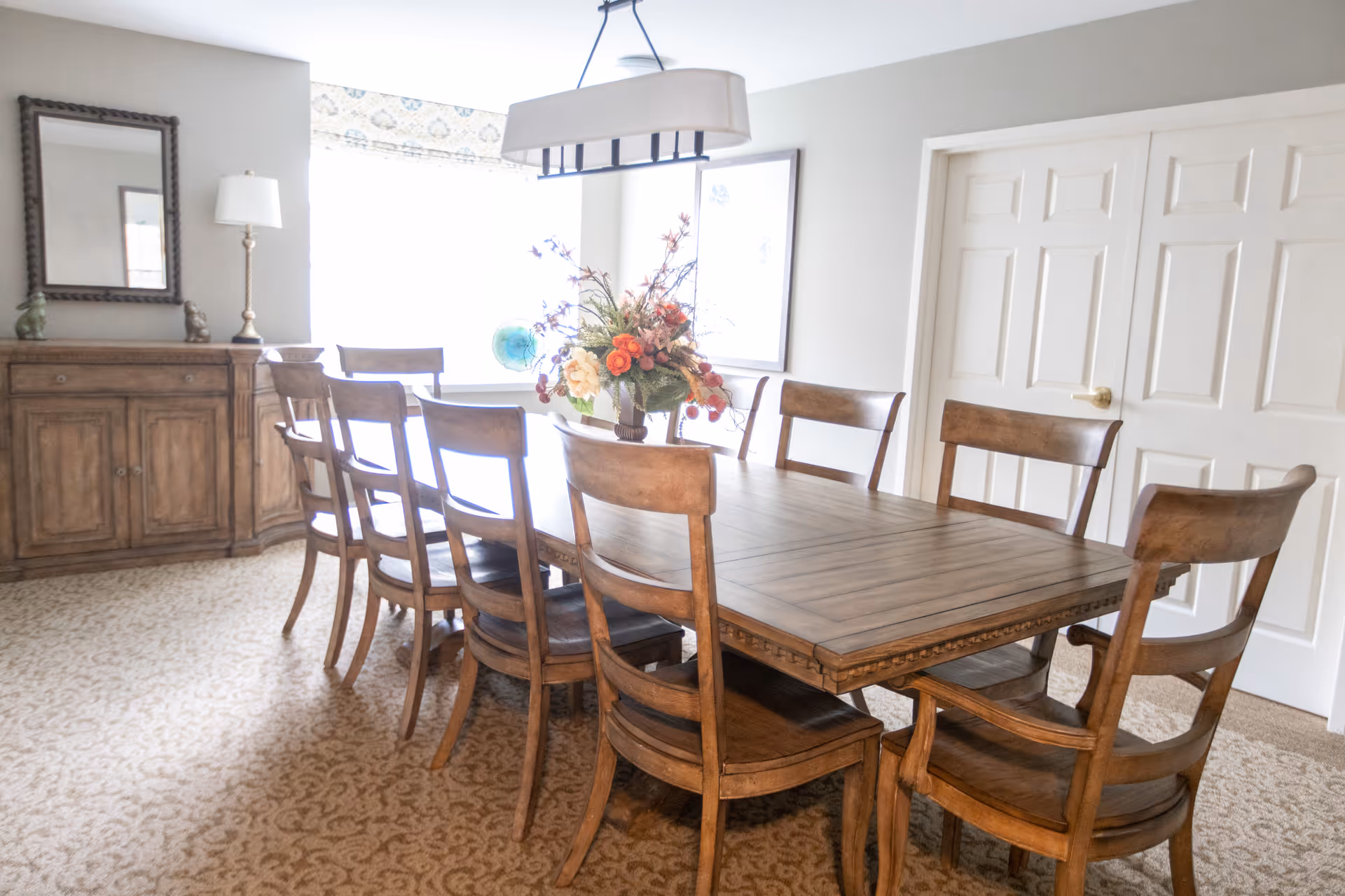 A bright dining room with a long wooden table surrounded by eight matching wooden chairs. A floral centerpiece is placed on the table. The room features a patterned carpet, a wooden sideboard with a lamp and decorative items, a large mirror on the wall, and a window with a patterned valance allowing natural light to fill the space. There are double white doors on one side of the room.
