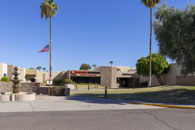 Exterior view of the Phoenix Mountain facility building with a clear blue sky, palm trees, a fountain, and an American flag on a pole. The building has a sign that reads 'Phoenix Mountain' and an entrance visible on the right side.