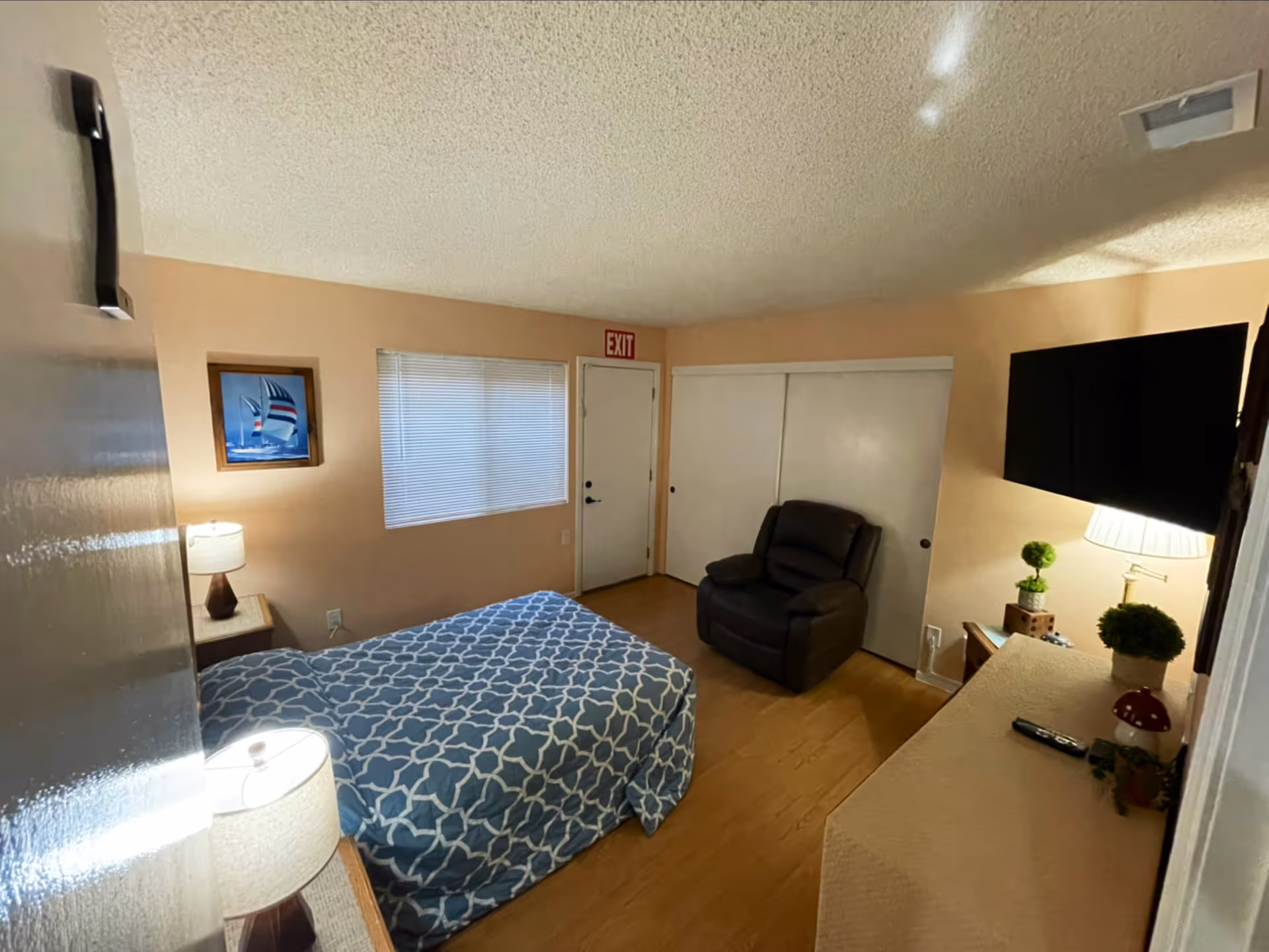 A cozy bedroom with a single bed covered in a blue and white patterned bedspread. There is a nightstand with a lamp next to the bed, a framed picture of sailboats on the wall, a window with closed blinds, a dark recliner chair, a dresser with a lamp and small plants, and a wall-mounted flat screen TV. The room has light-colored walls and wooden flooring.