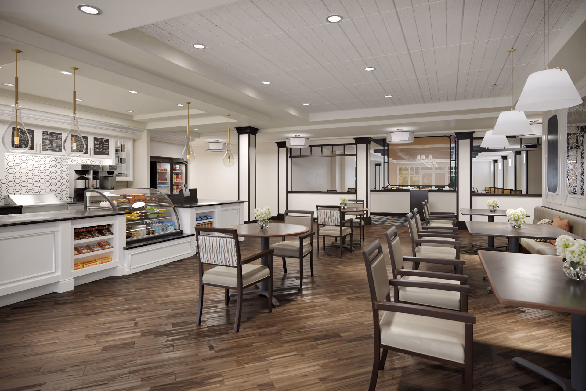 A bright and spacious dining area with wooden floors, multiple tables and chairs arranged neatly, and a counter with a display case containing snacks and beverages. The room features modern pendant lights, a coffee machine, and a refrigerator in the background. White walls and ceiling with recessed lighting create a clean and welcoming atmosphere.