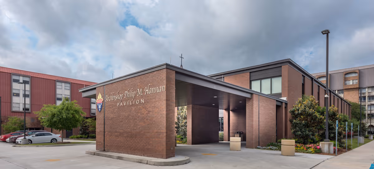 Exterior view of the Archbishop Philip M. Hannan Pavilion, a brick building with a covered entrance and a cross on top. There are parked cars, trees, and a cloudy sky in the background.