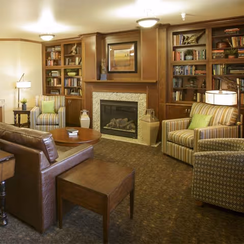 A cozy living room with a brown leather sofa, two striped armchairs with green cushions, a wooden coffee table, and built-in wooden bookshelves flanking a fireplace. The room is warmly lit with ceiling lights and a floor lamp.