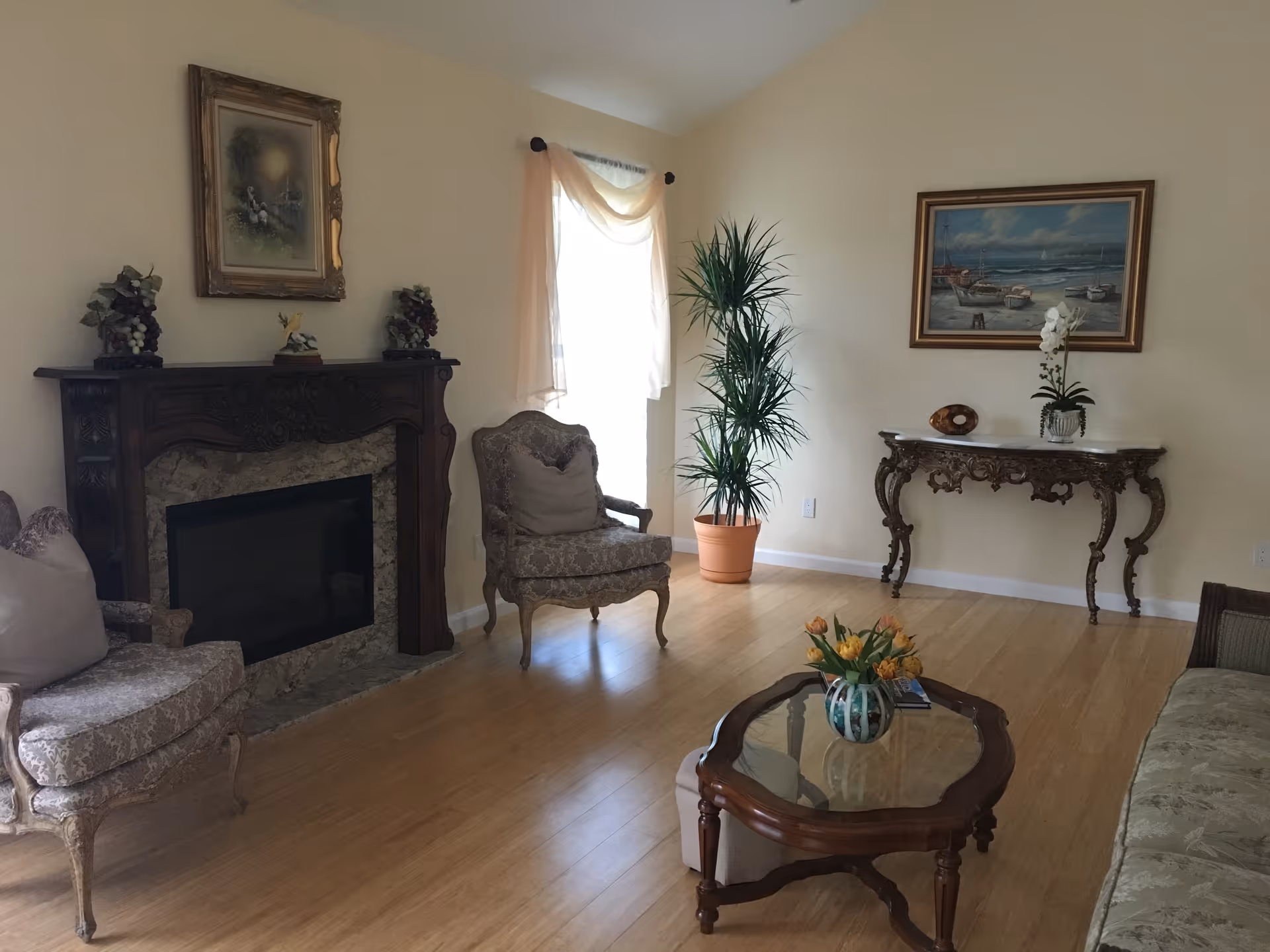 A cozy living room with vintage-style furniture including two armchairs and a sofa, a wooden coffee table with a vase of yellow flowers, a decorative fireplace with framed artwork above it, a tall potted plant near a window with sheer curtains, and an ornate console table with a flower arrangement and a decorative item on it.
