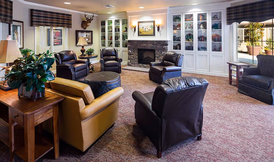 A cozy living room area in a memory care community featuring several leather armchairs arranged around a central ottoman. The room has a stone fireplace with a painting above it, built-in white cabinets with glass doors displaying decorative items, and large windows letting in natural light. There is a wooden side table with a lamp and a potted plant, and the carpet is a warm, patterned design.