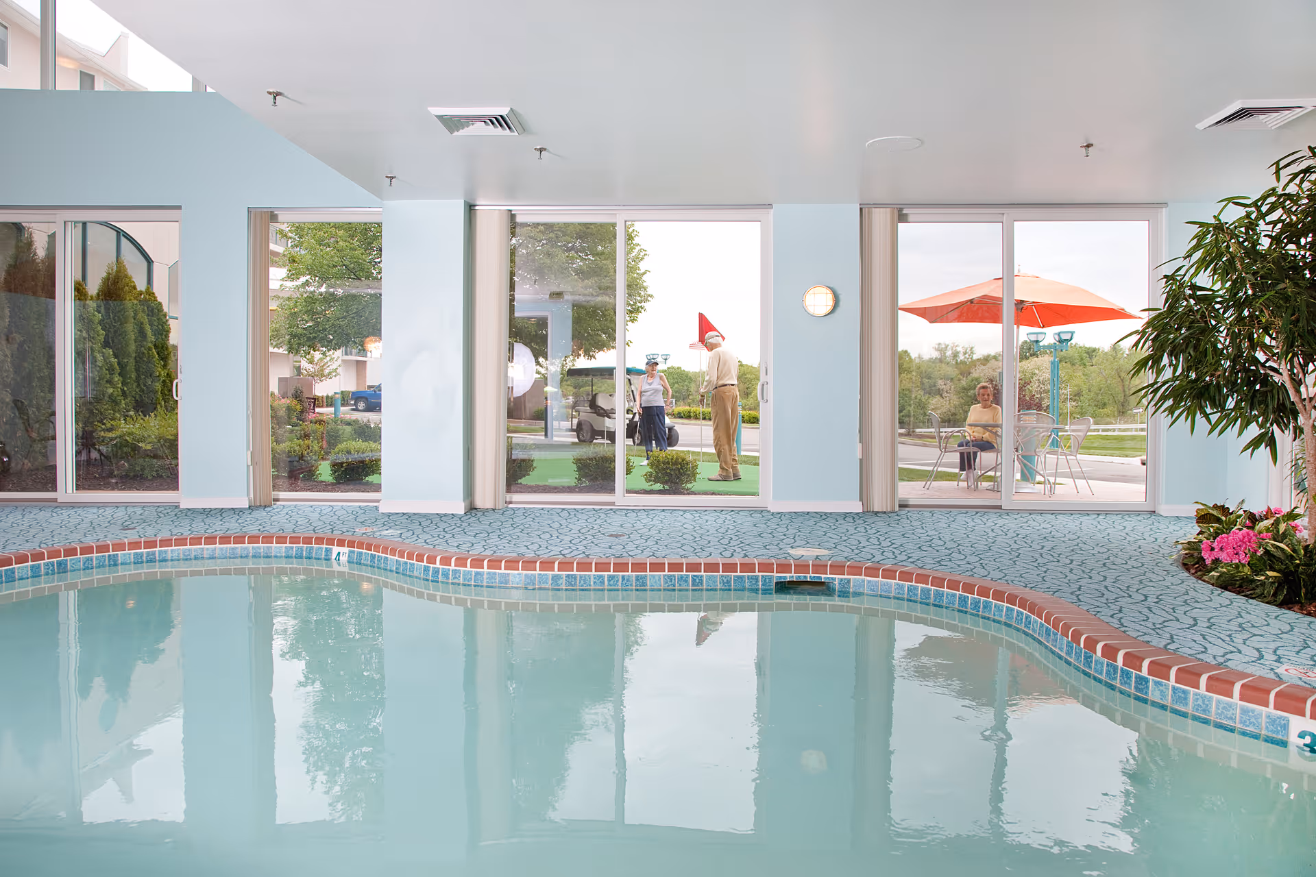 Interior pool area with a curved tiled swimming pool, patterned deck, and large floor-to-ceiling windows overlooking an outdoor patio with seating and people.