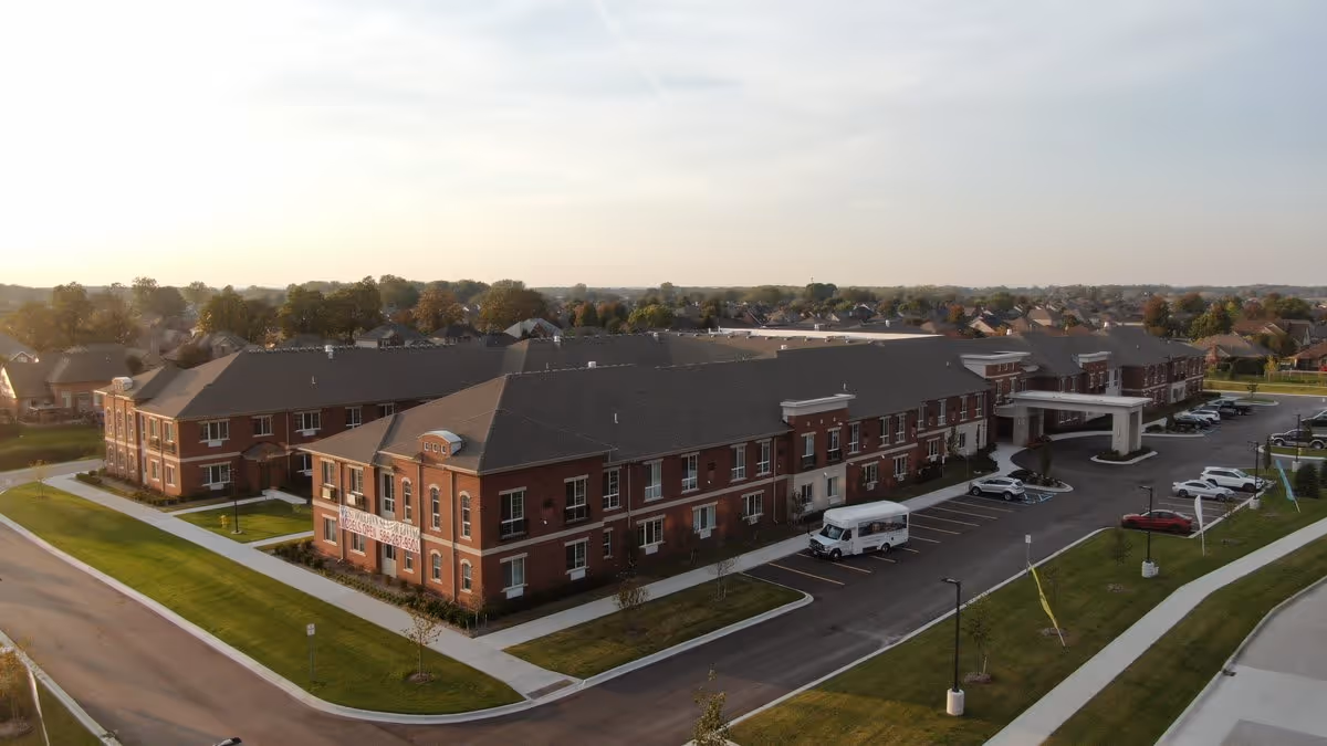 Aerial view of a large two-story brick senior living community building with a covered entrance, parking lot with several cars and a shuttle van, surrounded by green lawns and sidewalks, with a suburban neighborhood in the background under a partly cloudy sky.