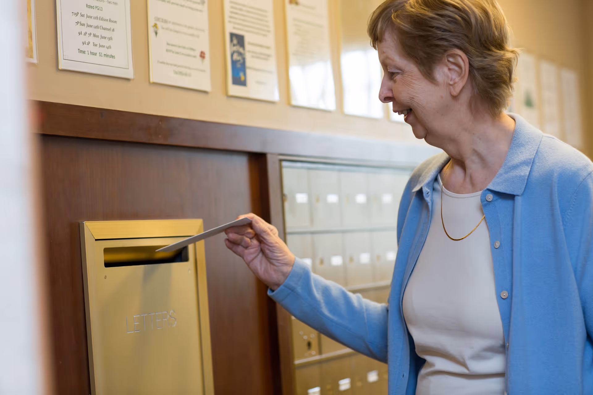 An elderly woman wearing a light blue cardigan and white top is smiling as she inserts a letter into a gold-colored mailbox slot labeled 'LETTERS' inside a building. Behind her, there are multiple small mailboxes and some posted notices on the wall.