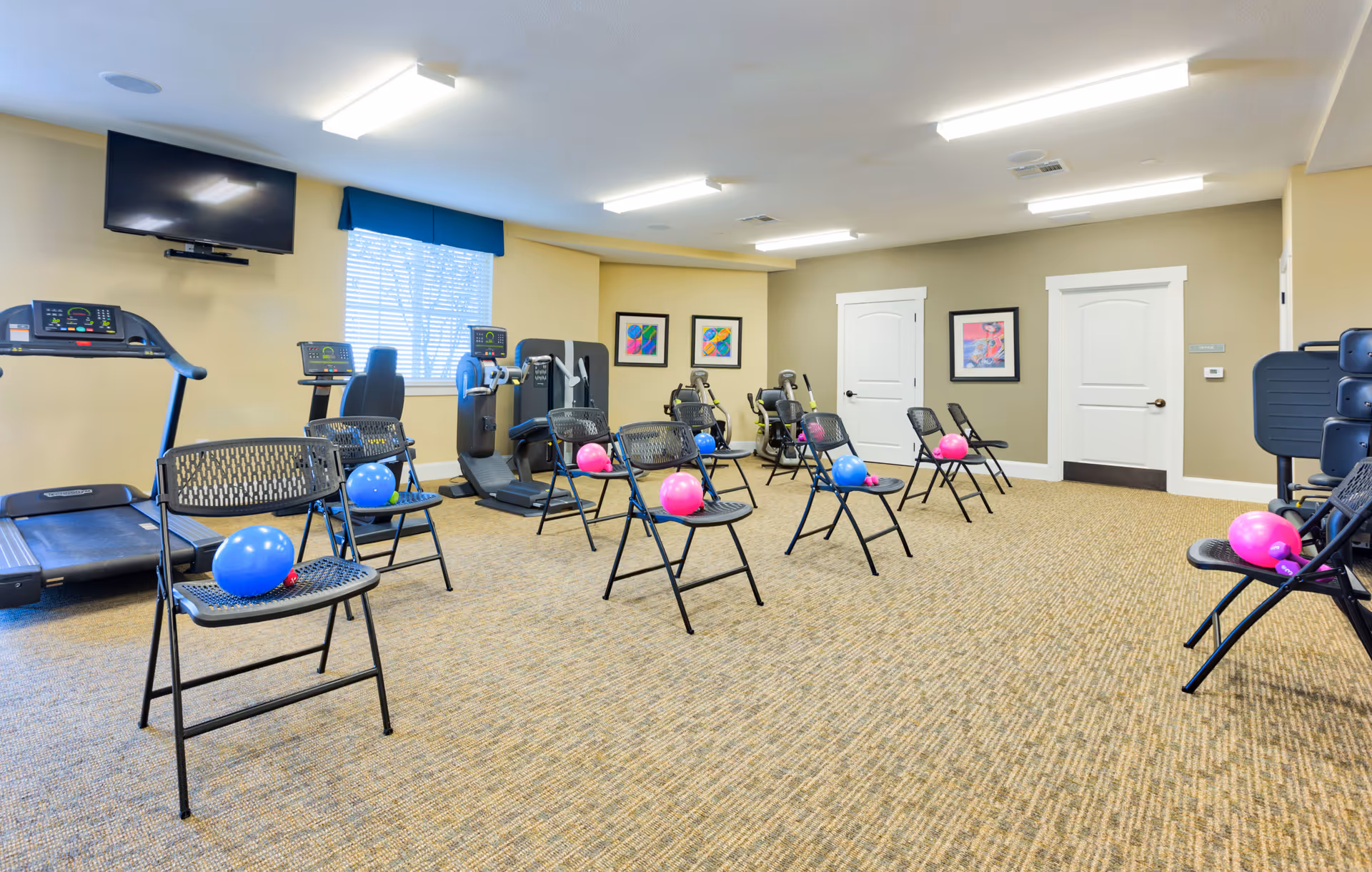 A fitness room with exercise equipment including treadmills and stationary bikes along the walls. Several black folding chairs are arranged in rows in the center of the room, each with a small pink or blue exercise ball placed on the seat. The room has beige walls, carpeted floor, fluorescent ceiling lights, and framed artwork on the walls. A window with blinds and a mounted flat-screen TV are visible on one wall.