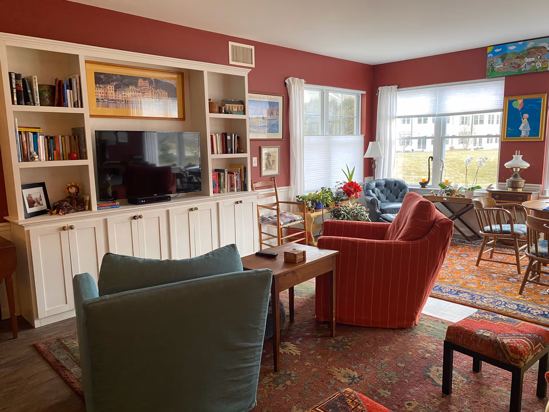 A cozy living room with red walls and large windows letting in natural light. The room features a built-in white bookshelf with a TV, various books, and framed pictures. There are two armchairs, one red and one blue, a wooden side table, several plants, and a round dining table with wooden chairs. Colorful rugs cover the floor, and framed artwork decorates the walls.