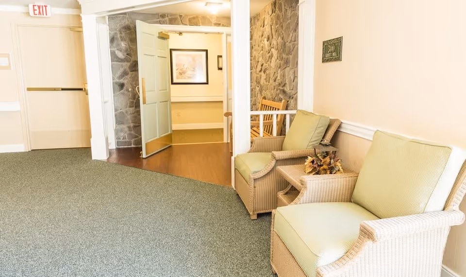 A cozy sitting area in a senior living facility hallway featuring two cushioned wicker armchairs with light green cushions and a small wicker side table with a decorative floral arrangement. The hallway has a green carpet, a stone accent wall, a wooden bench, and an open door leading to another room with framed artwork on the wall. An exit sign is visible above a closed door on the left.