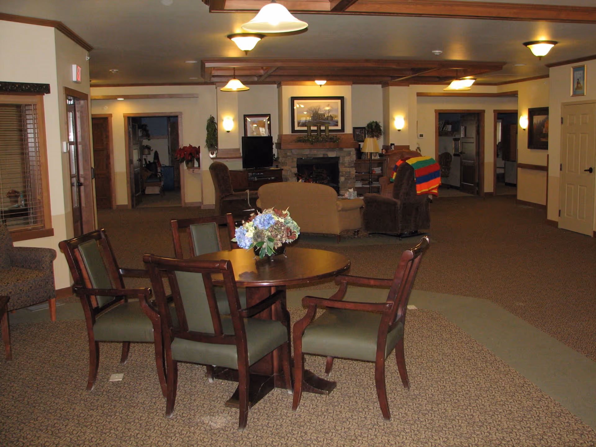 A cozy common area in a senior living facility featuring a round wooden table with four chairs and a floral centerpiece in the foreground. In the background, there is a seating area with armchairs and a loveseat arranged around a stone fireplace with a framed picture above it. The room has warm lighting with ceiling and wall fixtures, beige walls, and carpeted floors. Doorways and hallways lead to other parts of the facility.