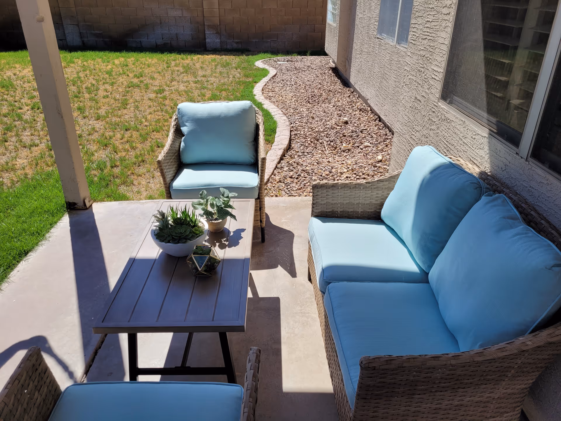 Patio with wicker seating and light blue cushions around a coffee table topped with potted succulents next to a small yard.
