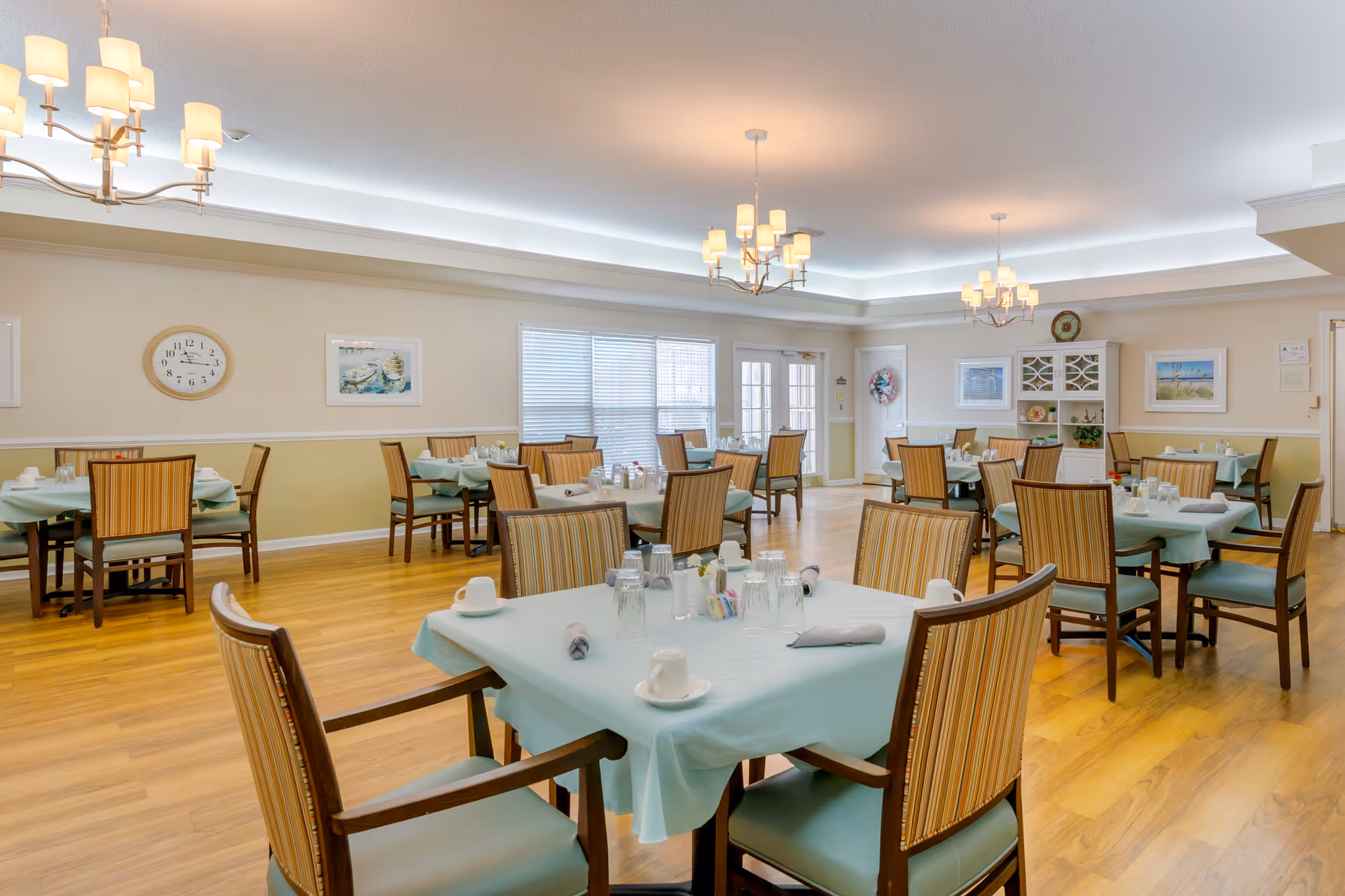A bright dining room with multiple tables covered in light blue tablecloths, each set with cups, glasses, napkins, and condiments. The room has wooden floors, beige walls with white trim, several framed pictures, a clock, and three modern chandeliers hanging from the ceiling. Large windows and glass doors allow natural light to fill the space.