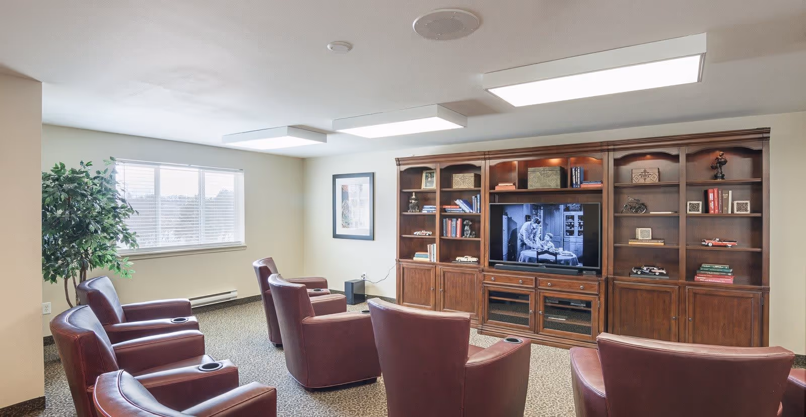 A cozy living room with six maroon leather armchairs arranged in two rows facing a large wooden entertainment center with shelves and cabinets. A black and white TV show is playing on the flat-screen television. There is a large window with blinds on the left wall and a green potted plant nearby. The room has beige walls and carpeted flooring with overhead rectangular light fixtures.