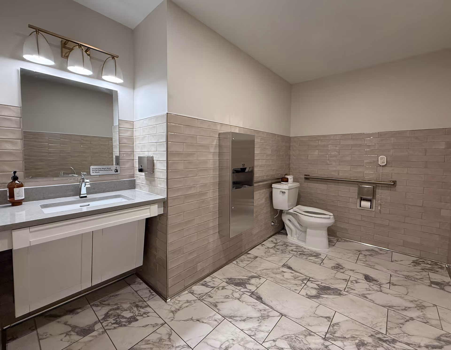 Spacious accessible restroom featuring a sink and mirror on the left and a toilet with a grab bar on the right, beige tiled walls and marble-pattern floor.