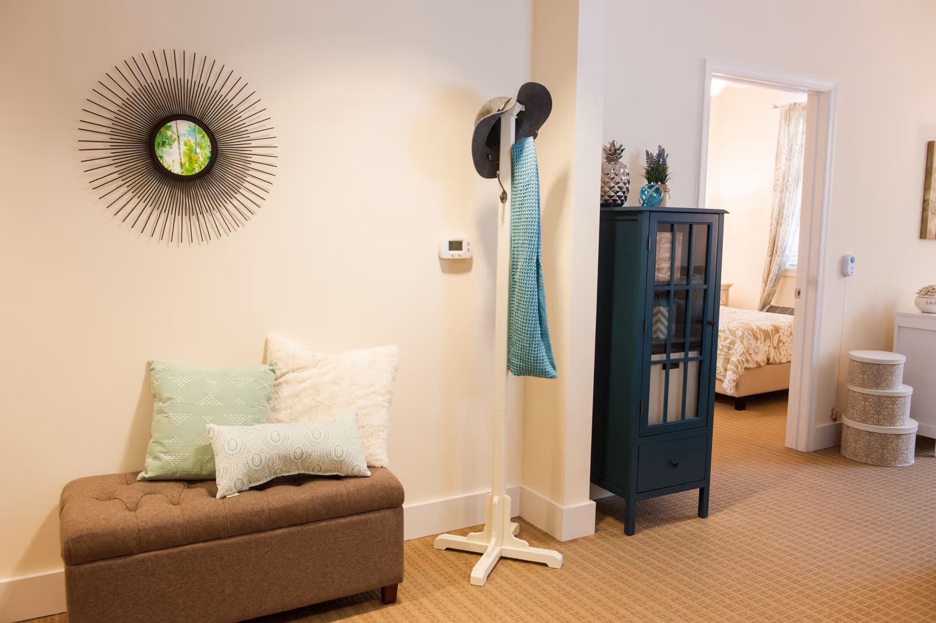 A cozy interior corner of a senior living facility featuring a brown cushioned bench with three decorative pillows, a white coat rack holding a gray hat and a blue scarf, a dark teal cabinet with decorative items on top, and a glimpse into a bedroom with a bed covered in a floral bedspread. The walls are light-colored and the floor is carpeted.